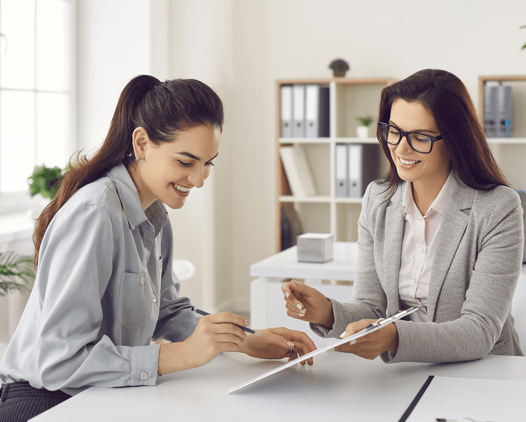 Two women in an office setting are discussing documents. One is smiling and writing on a clipboard while the other, wearing glasses, gestures towards the paper. Shelves and windows are in the background.
