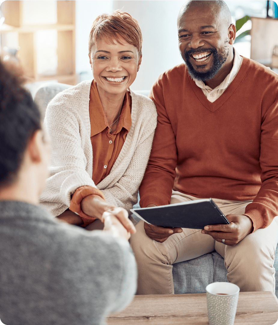 A couple sitting on a couch, smiling and shaking hands with a person in the foreground. The man holds a tablet. A coffee cup is on the table.