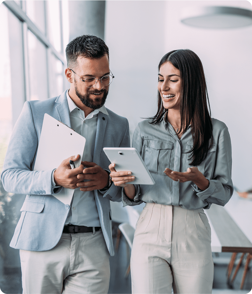 Two people in business attire stand in an office. One holds a clipboard, the other a tablet. They appear to be having a discussion.